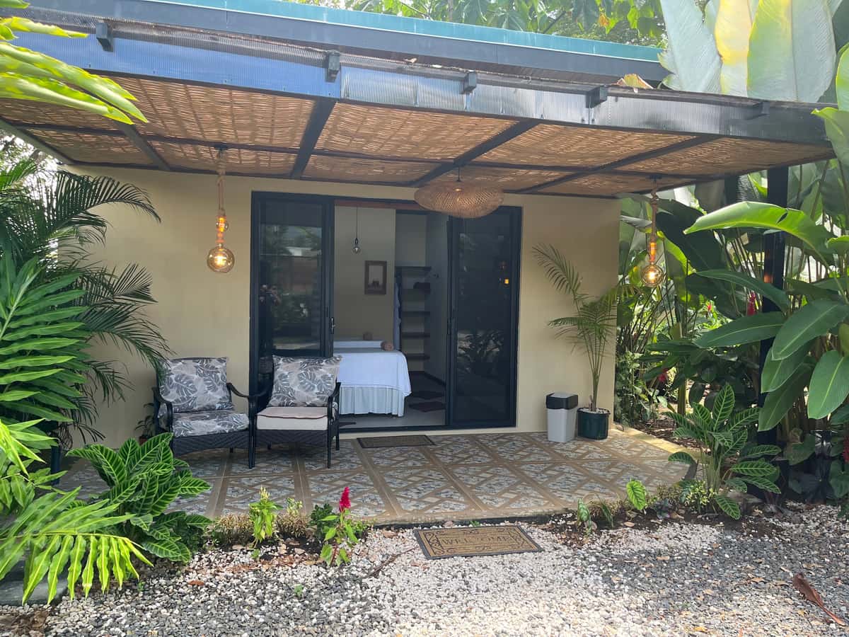 La entrada del bungalow de Reborn Spa bajo la pérgola, con dos sillones y la camilla visible por la puerta